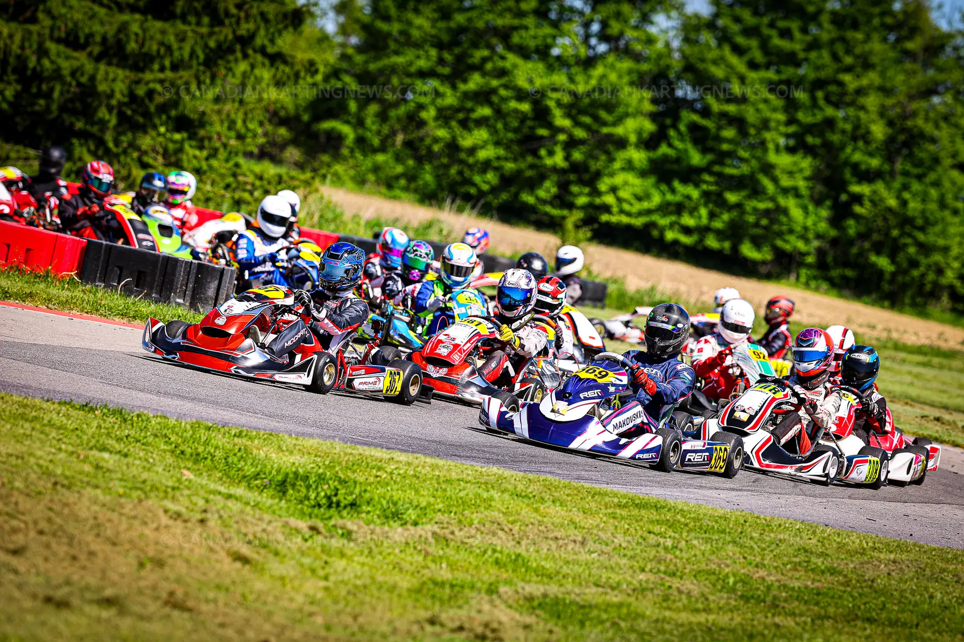 367OWENMCCARTHY369MAJORMAKOVSKIS26RMCONTR1CKN8922 Elementor Hosted Website Go-kart racers lean into a sharp turn on a track, colorful helmets and suits creating a rainbow of tones against a green infield and tires along the barrier edge behind them.