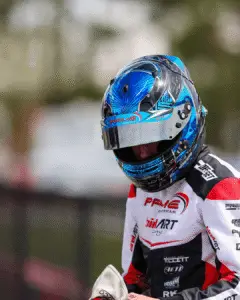 6 Elementor Hosted Website Elementor Hosted Website Motorsport racer wearing a blue patterned helmet and white-red racing suit with sponsor logos, preparing for a race.